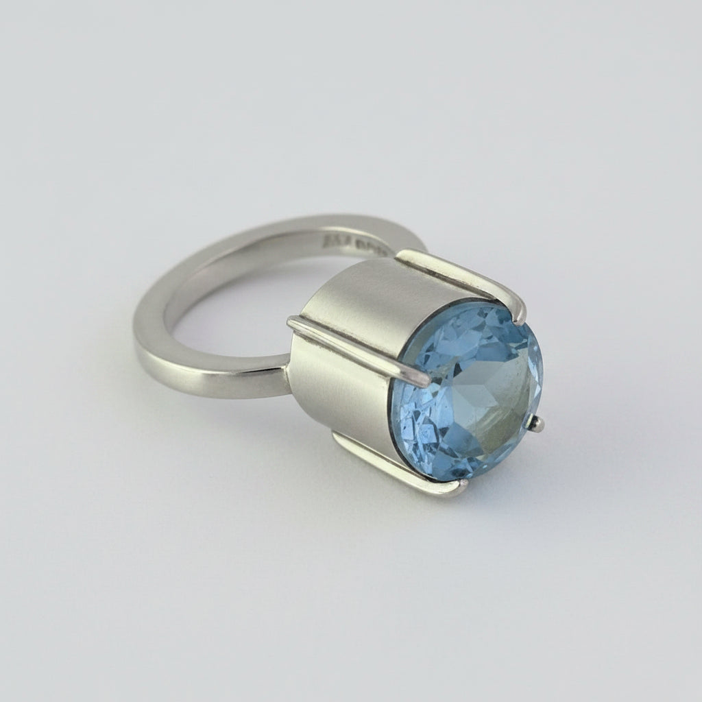 Silver Ring Set with 11mm Blue Topaz Gemstone Lying Flat on a White Background