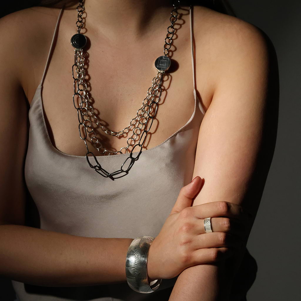 Torso of a girl wearing a silk silver top and silver jewellery by Antonello Figlia, a bangle, a chain necklace and a ring on a dark background