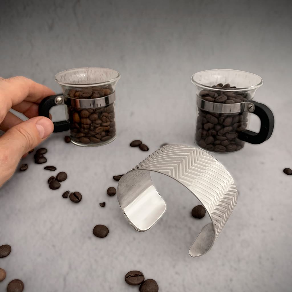 two coffee cups filled with coffee beans and a silver wide bracelet on a surface