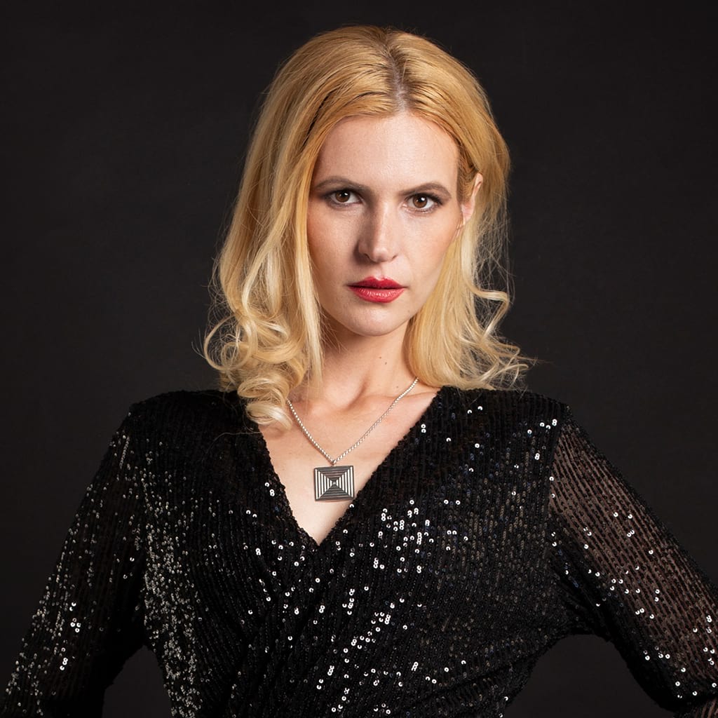 Blond model wearing a sparkling black dress and a square contemporary necklace on a dark background 
