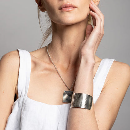 model touching her face with left arm on a grey background wearing a wide cuff bracelet and a square silver pendant